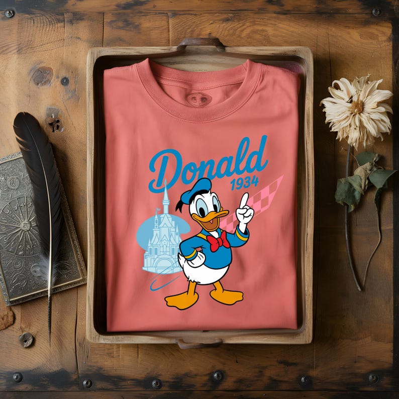 K&ouml;nnte beinhalten: Korallfarbenes T-Shirt mit einer Grafik von Donald Duck, der nach oben zeigt. Das Design beinhaltet das Wort "Donald" in blauer Schrift, das Jahr "1934" und eine Schlossillustration. Das Shirt wird in einem Holztablett pr&auml;sentiert.