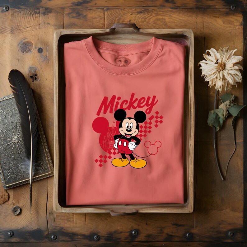 May include: A coral-colored t-shirt featuring a graphic of Mickey Mouse. The design includes the word "Mickey" in red script, a Mickey Mouse silhouette, and a checkered pattern. The shirt is displayed in a wooden tray.