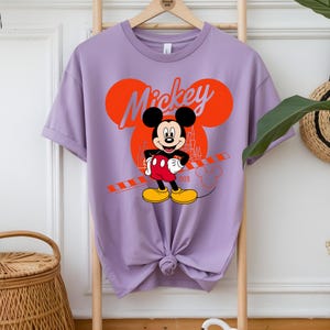 K&ouml;nnte beinhalten: Ein lilafarbenes T-Shirt mit einem Mickey Mouse-Motiv. Das Design zeigt Mickys Gesicht, das Wort "Mickey" in Orange und eine Schlossillustration. Das T-Shirt ist unten geknotet.