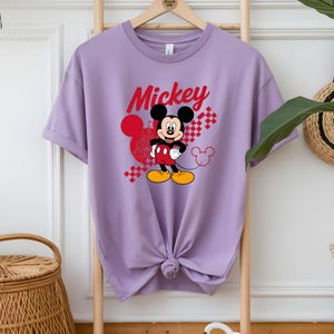 May include: A lilac-colored t-shirt featuring a graphic of Mickey Mouse. The word "Mickey" is written in red script above the character. Mickey Mouse is depicted in his classic outfit, with red shorts, yellow shoes, and white gloves.