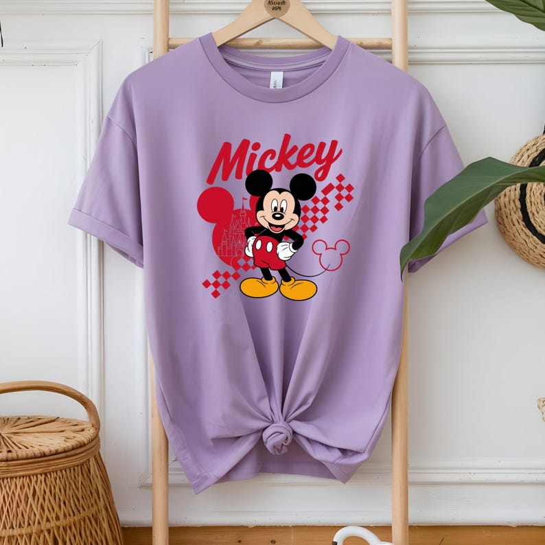 May include: A lilac-colored t-shirt featuring a graphic of Mickey Mouse. The word "Mickey" is written in red script above the character. Mickey Mouse is depicted in his classic outfit, with red shorts, yellow shoes, and white gloves.