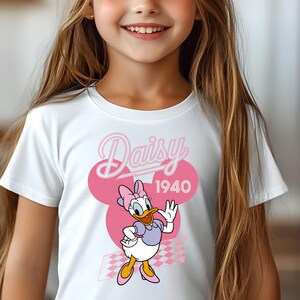 K&ouml;nnte beinhalten: Wei&szlig;es T-Shirt mit einem Daisy Duck-Motiv in Rosa und Lila, mit dem Text "Daisy 1940". Die Cartoonfigur winkt. Das Design umfasst rosa Kreise und ein Schachbrettmuster.
