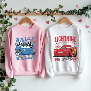 Puede incluir: Dos sudaderas de color rosa y blanco con temática de Cars. La sudadera rosa presenta un coche azul con el texto "Sally Cars" y la sudadera blanca presenta un coche rojo con el texto "Lightning McQueen".