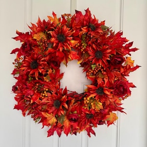 May include: A vibrant autumn wreath featuring red sunflowers, maple leaves, and berries. The wreath is composed of various shades of red, orange, and yellow, creating a warm and inviting seasonal decoration. The wreath is hanging on a white door.