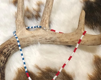 May include: A beaded necklace with a patriotic color scheme. The necklace features small, round beads in red, white, and blue, strung together to create a striking visual pattern. The necklace is displayed on a deer antler against a backdrop of faux fur.