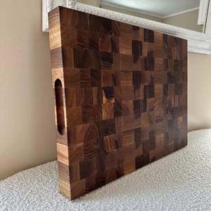 May include: A large, rectangular end-grain cutting board made of dark brown wood. The board features a mosaic-like pattern created by the end-grain construction. It has a built-in handle on one side for easy carrying. The board is designed for kitchen use.