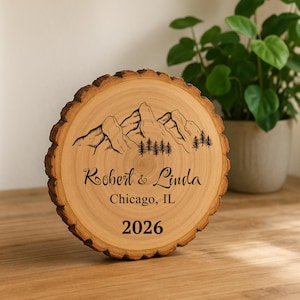 May include: A round wooden plaque with a natural bark edge. The plaque features an engraved mountain scene with trees, the names "Robert & Linda", the city "Chicago, IL", and the year "2026".