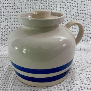 May include: A cream-colored ceramic crock with a matching lid and a single side handle. The body of the vessel features two distinct horizontal blue stripes encircling the lower portion, providing a clear contrast against the smooth, glossy finish.