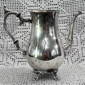 May include: A silver-colored, antique coffee pot with an ornate handle and spout. The pot has a bulbous body with a decorative pattern near the base and rests on three small feet. The handle is curved and detailed, adding to the vintage aesthetic.