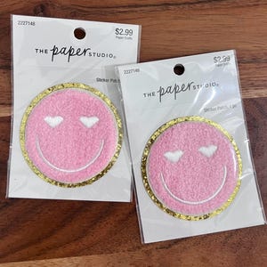 May include: Two pink smiley face sticker patches with heart-shaped eyes and a gold glitter border. Each patch is packaged in a clear plastic sleeve with the text "THE paper STUDIO" and "Sticker Patch".