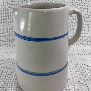 May include: A cylindrical ceramic pitcher with a light beige, speckled finish and a smooth glaze. The vessel features two horizontal, hand-painted cobalt blue stripes encircling the body, a curved handle on the right side, and a small pouring spout.