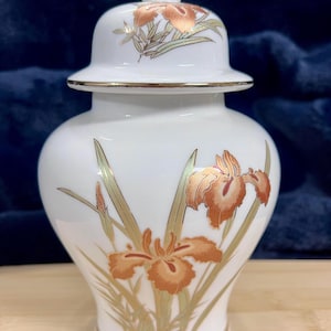May include: White ceramic jar with a lid, adorned with gold trim and iris flower designs in shades of orange and gold. The jar has a rounded body and a lid with a gold finial. The iris flowers are painted on the body and lid.