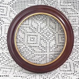 May include: A circular picture frame with a smooth, dark mahogany-colored finish and a thin, metallic gold-toned inner rim. The frame features a rounded, convex profile that curves outward, creating a simple and clean border for a circular image.