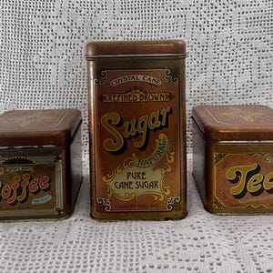 May include: A set of three rectangular metal canisters with brown and gold vintage-style labels. The center canister is tall and labeled Sugar, while the two smaller side canisters are labeled Coffee and Tea, all featuring decorative borders.