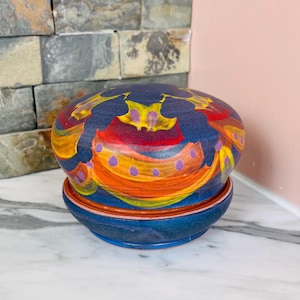 May include: A hand-painted ceramic pot with a matching saucer. The pot is a deep blue with vibrant abstract designs in yellow, red, orange, and purple. The saucer is a similar blue with a thin orange stripe. The pot is sitting on a white marble surface.