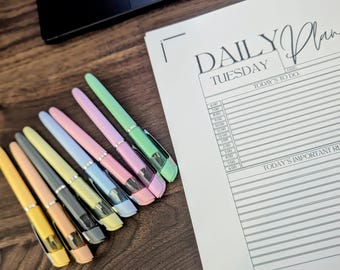 Op de afbeelding: Een verzameling kleurrijke pennen in tinten geel, oranje, grijs, blauw, roze en groen, diagonaal gerangschikt op een houten oppervlak. Een "Daily Plan" notitieblok is zichtbaar op de achtergrond, met de dag "Tuesday" erop gedrukt.