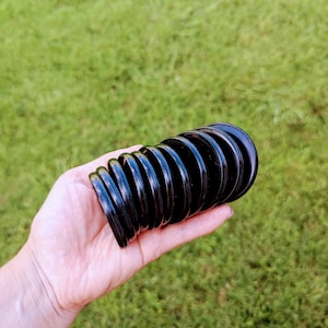 May include: A black, accordion-style object with a series of circular, ridged segments. The object is held in a hand against a backdrop of green grass. The segments appear to be made of a glossy material.