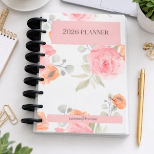 May include: A 2026 planner with a floral design featuring pink roses and orange accents. The planner has a black binding and the text "2026 PLANNER" on the cover. A gold pen rests beside the planner.