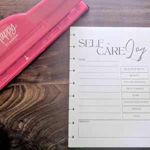 May include: A pink Happy Planner hole punch and a white Self-Care Joy planner sheet. The planner sheet has sections for date, health, beauty, fun, home, personal development, spiritual, social, and thoughts.