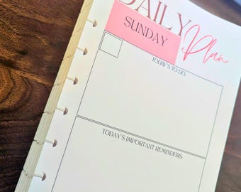 May include: A white planner page with the words "DAILY Plan" in pink script. The page includes sections for "TODAY'S TO DO" and "TODAY'S IMPORTANT REMINDERS". The planner is open to the "SUNDAY" page.