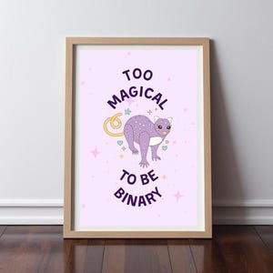 Peut inclure: Une impression encadrée avec un animal de dessin animé violet et blanc sur un fond rose. Le texte sur l'impression est "Too Magical To Be Binary".