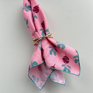 May include: Pink cloth napkin with a radish pattern, held by a gold-colored napkin ring. The napkin has a teal green trim and a repeating pattern of purple radishes and teal green leaves.