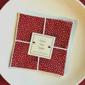 May include: A set of four red cocktail napkins with white polka dots, tied together with white string, and a tag that reads "Farm Girl Table". The napkins are presented on a white plate.