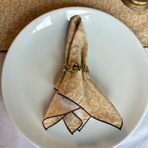 May include: A folded tan napkin with a white pumpkin pattern, set on a white plate. The napkin is held by a gold leaf-shaped napkin ring. The napkin has a dark brown border. The background includes a matching pumpkin-patterned tablecloth.