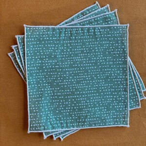 May include: A stack of square, green fabric napkins with a white dotted pattern and white stitched edges. The napkins are arranged on a brown surface.