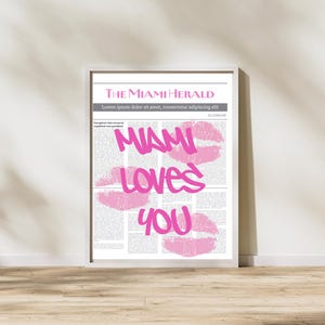 May include: Framed print featuring a newspaper-style design with the headline "THE MIAMI HERALD." The text "MIAMI LOVES YOU" is displayed in pink, along with pink lip prints. The print is in a white frame.