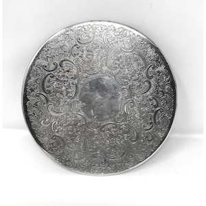 May include: A circular silver-colored metal tray featuring an intricate embossed pattern of floral motifs and scrolling vines surrounding a plain, smooth, unadorned center. The metallic surface has a reflective finish with detailed engravings.