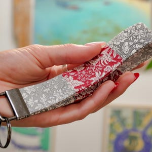 May include: A fabric wristlet key fob with a silver-tone metal clasp and key ring. The key fob features a patchwork design with floral patterns in red, white, and gray. The key fob is held in a hand.