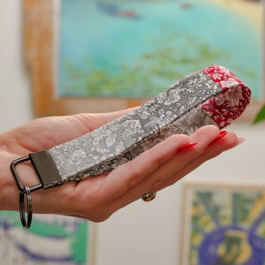 May include: A fabric keychain with a floral pattern in gray and white, accented with a red floral end piece. The keychain has a black metal ring and hardware.