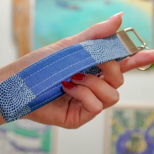 May include: A blue and white patterned wristlet keychain. The wristlet is made of fabric with a geometric design and a solid blue center. It has a metal clasp for attaching keys.