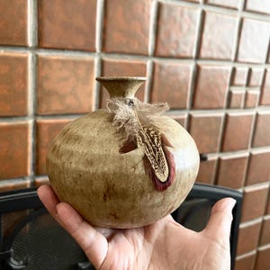 May include: A small, round, beige ceramic vase with a narrow neck and a decorative feather attached. The feather has brown and burgundy markings. The vase is held in a hand, with a brick-like background.
