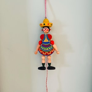 May include: A colorful wooden pull-string toy in the shape of a person wearing a yellow sombrero and a patterned dress. The toy has red accents and black shoes, hanging from a red and white string.