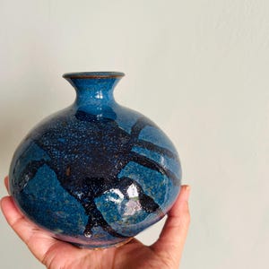 May include: A round, blue ceramic vase with a narrow neck. The vase features a speckled texture and dark blue abstract design. The vase is held in a hand, showcasing its size and shape. The vase is a decorative item.