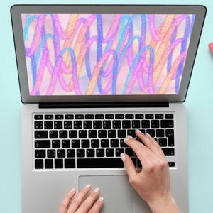 May include: A person is typing on a silver laptop with a colorful abstract design on the screen. The laptop is on a light blue surface with a pink notebook and a pair of glasses.