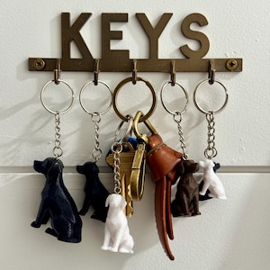 May include: A bronze metal key holder with the word "KEYS" across the top. The key holder has five hooks with keychains attached. The keychains feature small dog figurines in black, white, and brown.