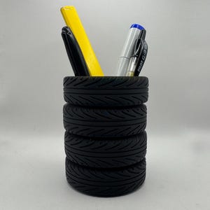 May include: A black pen holder made from stacked miniature tires. The pen holder contains a yellow highlighter, a black pen, and a silver pen with a blue cap. The tires have detailed tread patterns.