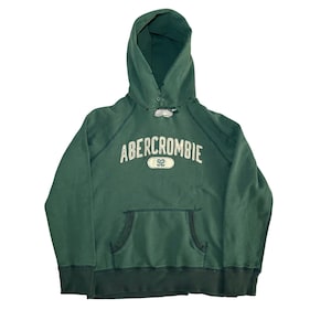May include: A green Abercrombie hoodie with a drawstring hood and a kangaroo pocket. The word "Abercrombie" is arched across the chest in white lettering, with "92" below it. The hoodie is made of a soft, worn material.