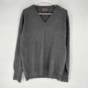 May include: A gray V-neck sweater made of cashmere. The sweater is hanging on a white hanger. The label on the sweater reads "100% Cashmere". The sweater has long sleeves and a classic design.