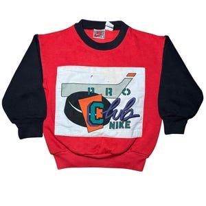 May include: Red and black vintage Nike sweatshirt with a hockey-themed graphic. The sweatshirt features a red body, black sleeves, and a white patch with a hockey stick, puck, and the words "Club Nike". The Nike logo is visible on the collar.
