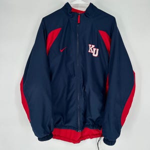 May include: Navy blue zip-up jacket with red accents and a red interior. Features the Nike logo and the letters "KU" in white. The jacket has a full zipper and elastic cuffs.