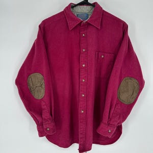 May include: A long-sleeved, button-down shirt in a deep red hue, featuring brown elbow patches and a chest pocket. The shirt has a collared neckline and buttoned cuffs. The shirt is hanging on a black hanger.