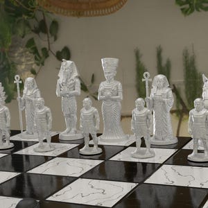 Könnte beinhalten: Ein weißes Schachspiel mit ägyptischem Thema. Die Figuren umfassen Pharaonen, Königinnen und andere Figuren, die auf einem schwarz-weißen Schachbrett angeordnet sind. Die Schachfiguren sind aufwendig detailliert.