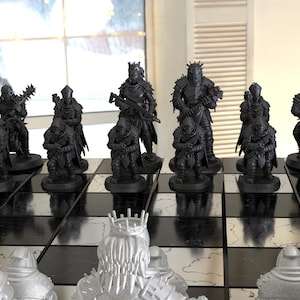 Puede incluir: Un juego de ajedrez negro y plateado con piezas detalladas de temática fantástica. Las piezas negras presentan figuras blindadas y máquinas de guerra, mientras que las piezas plateadas tienen un diseño similar. El tablero es blanco y negro.