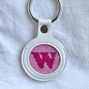 May include: White leather keychain with a pink stitched design featuring the letter "W" in a darker pink. The keychain has a silver metal ring for attaching to keys or bags. The design is circular.