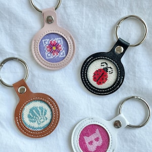 Peut inclure: Quatre porte-clés en cuir avec des anneaux métalliques. Chaque porte-clés présente un motif cousu : une fleur, une coccinelle, un coquillage et la lettre "B". Les porte-clés sont roses, noirs, marron et blancs.