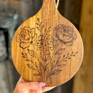May include: A round wooden cutting board with a handle and a floral design. The board is made of light brown wood and features a laser-etched bouquet of flowers, including roses and peonies. A natural rope is attached to the handle.